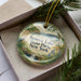 A decorative ornament in a box, featuring a scenic landscape with a lake, trees, and a bird.