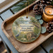 A wooden tray with a decorative ornament featuring a scenic landscape of Sesquicentennial State Park, South Carolina, and pine cones.