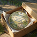 A decorative ornament in a brown box, featuring a scenic landscape with a house, trees, and a body of water.