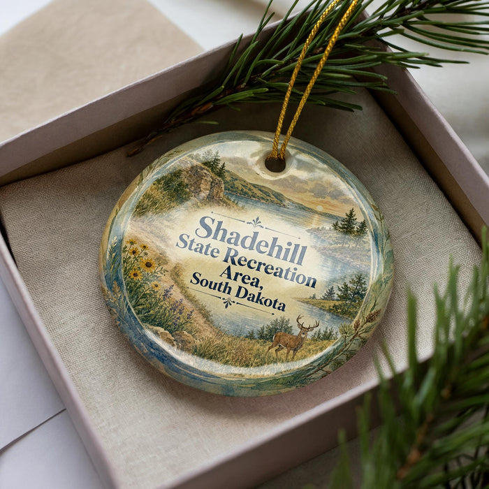 A decorative ornament with a scenic landscape, featuring a deer, mountains, and a body of water, is displayed in a box with pine branches.