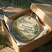 A round ornament in a brown box, featuring a scenic landscape with a lake and mountains, is displayed on a wooden surface.
