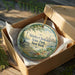 A decorative ornament in a cardboard box, featuring a scenic image of Shirley Chisholm State Park in New York.