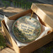 A round ornament in a brown box, featuring a scenic landscape with a lighthouse and trees, with the text "Shore Acres State Park, Oregon" printed on it.