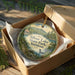 A round ornament in a box, featuring a scenic landscape with mountains, trees, and a lake, with the text "Silver Lake State Park, Vermont" printed on it.