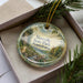A decorative ornament with a scenic landscape of Sizersville State Park, Pennsylvania, displayed in a box with pine branches.