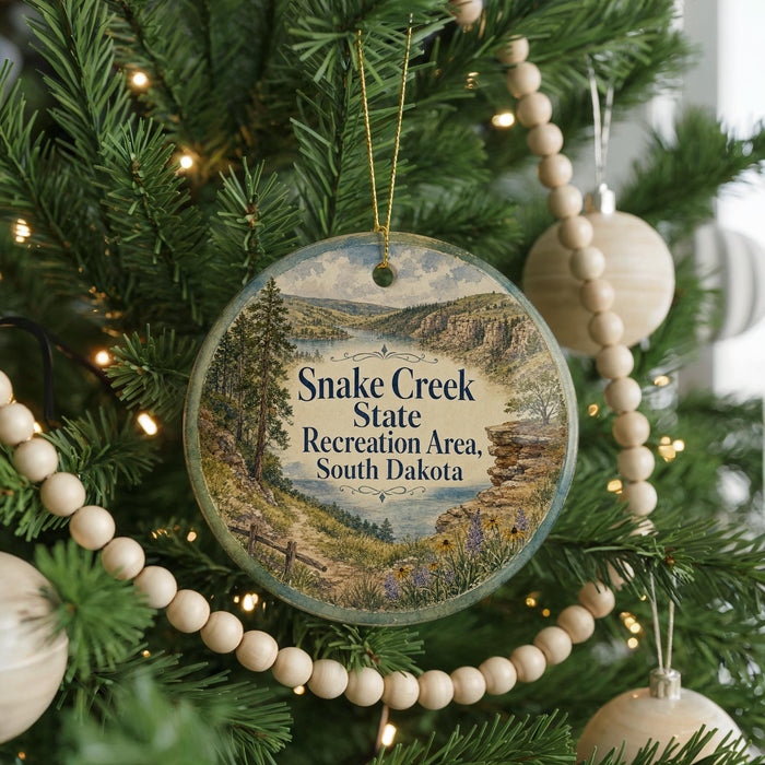A round ornament hanging on a Christmas tree, featuring a scenic landscape of Snake Creek State Recreation Area in South Dakota.