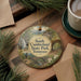 A decorative ornament with a scenic image of a park, featuring a waterfall, a bridge, and a person walking, is displayed on a wooden surface alongside a cup of coffee and some pine branches.