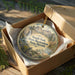 A round ornament in a brown box, featuring a scenic landscape with mountains, trees, and a waterfall.