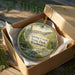 A decorative ornament in a wooden box, featuring a scenic landscape with a river, trees, and mountains.