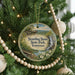 A round ornament hanging on a Christmas tree, featuring a scenic landscape with a waterfall and a bridge.