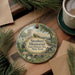 A decorative ornament featuring a statue of a man in historical attire, surrounded by a scenic landscape, placed on a wooden surface alongside a cup of coffee and some papers.