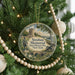 A round ornament hanging on a Christmas tree, featuring a scenic landscape with a monument and a bridge.
