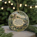 A round ornament with a painting of a monument and a cannon, surrounded by greenery and a string of lights.