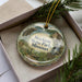 A decorative ornament in a box, featuring a scenic landscape with a river, mountains, and buildings.