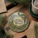 A decorative ornament with a scenic image of Stockton State Park, Missouri, featuring a waterfall, a cabin, and a path, is displayed on a wooden surface alongside a cup of coffee and some pine branches.