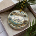 A decorative ornament in a box, featuring a scenic landscape with mountains, water, and wildlife.