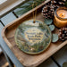 A decorative ornament featuring a scenic lake landscape with a boat, pine cones, and a candle on a wooden tray.