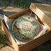A round ornament in a brown box, featuring a scenic landscape with a bird and pine trees, is displayed on a wooden surface.