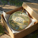 A decorative ornament in a box, featuring a scenic landscape with a house, bridge, and flowers, with the text "Sully Creek State Park, North Dakota" printed on it.