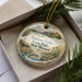 A decorative ornament in a box, featuring a scenic landscape with a lake, mountains, and a cabin.