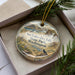 A decorative ornament with a landscape scene featuring a bird, placed in a box with pine branches.