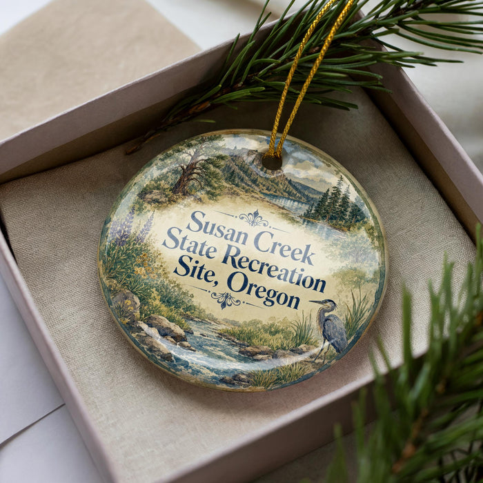 A decorative ornament in a box, featuring a scenic landscape with a river, mountains, and pine trees.