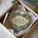 A round ornament with a scenic landscape and a bird, placed in a box with a white tissue paper lining.