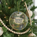 A round ornament hanging on a Christmas tree, featuring a scenic landscape with a river and trees.