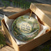 A round ornament in a box, featuring a scenic landscape with a waterfall and trees, with the text "TO Fuller State Park, Tennessee" printed on it.