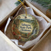 A decorative ornament in a box, featuring a scenic landscape with mountains, trees, and a waterfall.