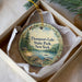 A round ornament with a scenic image of a lake and pine trees, hanging from a string inside a box.
