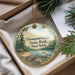 A decorative ornament with a scenic lake and forest landscape, featuring pine trees and a cabin, is displayed in a box with pine branches.