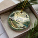 A decorative ornament in a box, featuring a scenic landscape with pine trees, a lake, and a bird in flight.