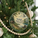 A round ornament hanging on a Christmas tree, featuring a scenic landscape with a cabin, lake, and mountains.