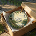 A circular ornament in a wooden box, featuring a scenic landscape with a lake, mountains, and a bird in flight.
