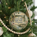 A round ornament hanging on a Christmas tree, featuring a scenic landscape with a lighthouse and a body of water.
