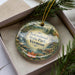 A decorative ornament with a scenic landscape of Vermont State Park, featuring a waterfall and pine trees, is displayed in a box with pine branches.