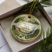 A decorative ornament with a natural landscape scene, featuring a forest, waterfall, and flowers, is displayed in a box with pine branches.