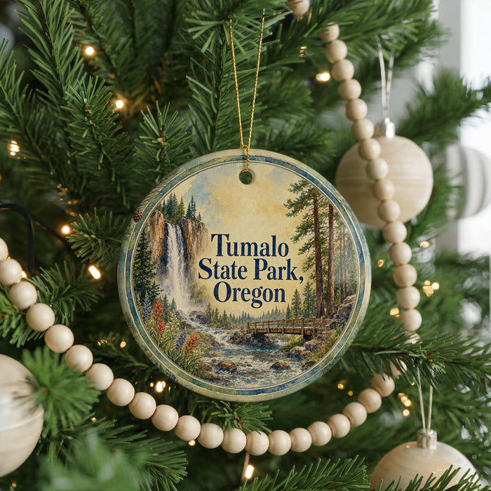 A round ornament hanging on a Christmas tree, featuring a scenic image of a waterfall surrounded by pine trees and a bridge.