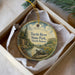 A round ornament with a painting of a turtle in a pond, surrounded by trees and mountains, placed in a box with a white tissue paper lining.
