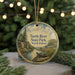 A round ornament with a scenic landscape of a river, pine trees, and a turtle resting on a rock, hanging from a string.