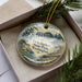 A decorative ornament in a box, featuring a scenic landscape with a lake, mountains, and pine trees.