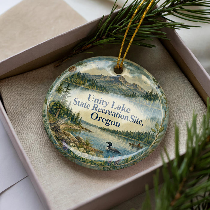 A decorative ornament in a box, featuring a scenic landscape with mountains, pine trees, and a lake.