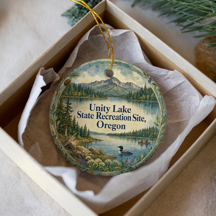 A decorative ornament in a white box, featuring a scenic landscape with a lake, mountains, and pine trees.