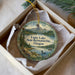 A decorative ornament in a white box, featuring a scenic landscape with a lake, mountains, and pine trees.