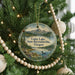 A round ornament hanging on a Christmas tree, featuring a scenic landscape with mountains, a lake, and pine trees.
