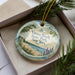 A decorative ornament with a beach scene and text, placed in a box with pine branches.