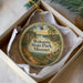 An oval-shaped ornament with a painting of a scenic landscape, featuring a path, flowers, and a house, is displayed in a box.