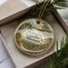 A decorative ornament in a box, featuring a scenic landscape with a river, mountains, and flowers.
