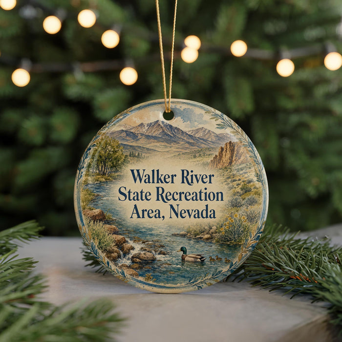 A round ornament with a scenic landscape of mountains, trees, and a river, featuring a duck swimming in the water. The ornament is hanging from a string and is placed on a surface with pine branches.